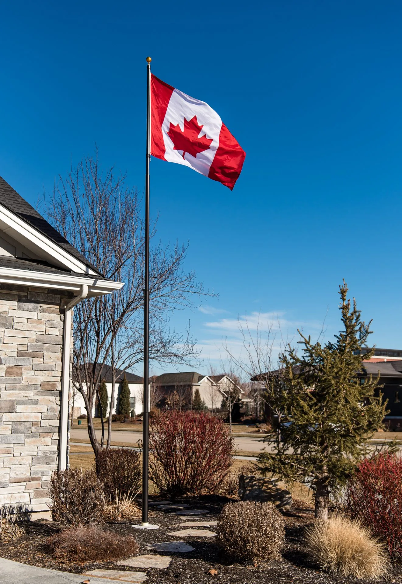 A 20 foot flagpole with a dark bronze finish provided by The Flag Shop. It is installed outside a house and is flying the Canadian flag.