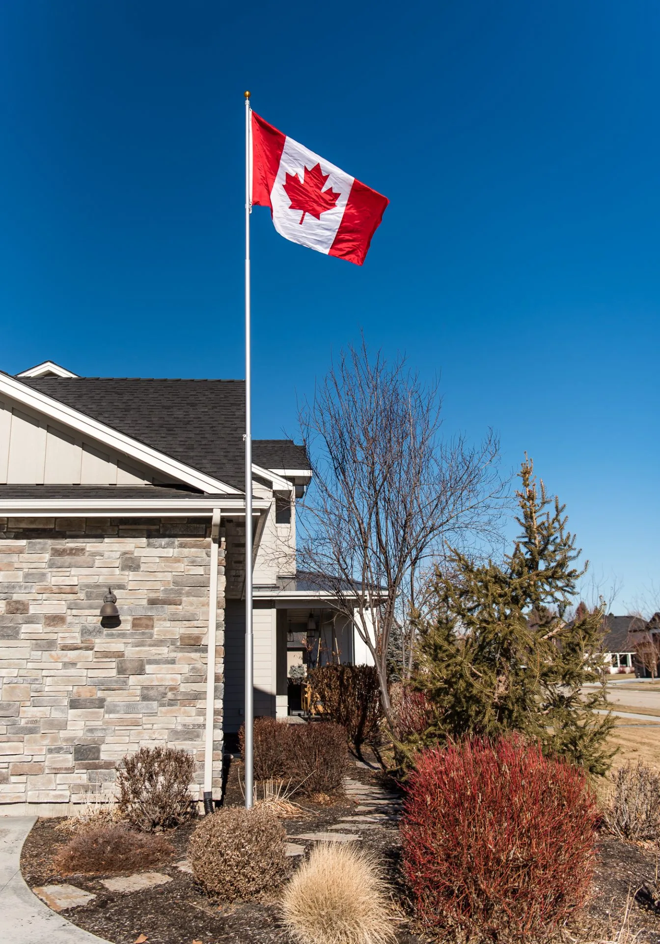 A 25 foot flagpole with a silver anodized finish provided by The Flag Shop. It is installed outside a house and is flying the Canadian flag.
